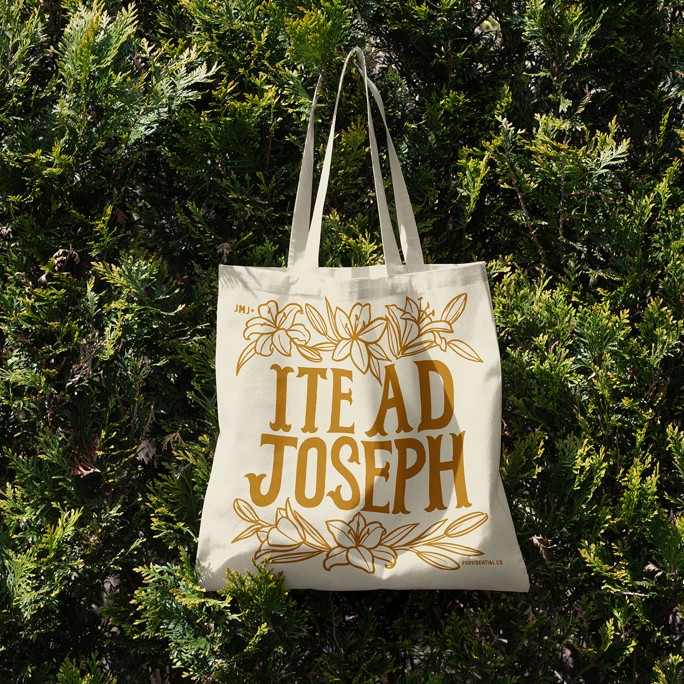 Tote bag with 'ITE AD JOSEPH' text and floral designs against a green bush background